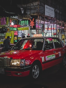A red taxi is parked on a bustling city street lined with brightly lit neon signs. Various signs in multiple languages advertise currency exchange and sales. Scaffolding is visible on a nearby building, and a few pedestrians are walking.