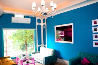 A modern living room with vibrant decor features a blue accent wall adorned with abstract art pieces. Two sofas in green shades are arranged around a glass coffee table with a bright pink metal frame. A white ceiling chandelier enhances the space, and natural light pours in through a large window displaying a view of green foliage. An air conditioning unit is mounted on the wall above the window.