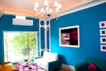 A modern living room with vibrant decor features a blue accent wall adorned with abstract art pieces. Two sofas in green shades are arranged around a glass coffee table with a bright pink metal frame. A white ceiling chandelier enhances the space, and natural light pours in through a large window displaying a view of green foliage. An air conditioning unit is mounted on the wall above the window.