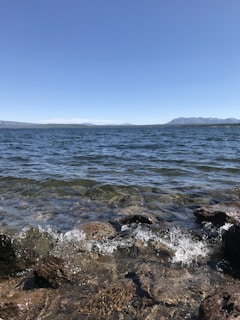 Clear blue lake water gently splashes against rugged rocks along the shore. In the distance, mountain ranges loom under a wide, cloudless sky. The water is transparent near the rocks, revealing pebbles underneath.