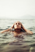 A person is peacefully floating in water with eyes closed and hands gently holding their head. The scene conveys a sense of calm and relaxation with serene water and muted background tones.