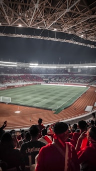 A large stadium filled with spectators, predominantly dressed in red, overlooks a well-lit football field with distinct grass patterns. The stadium's architecture features an intricate roof design with exposed beams. The field is surrounded by a running track, and the atmosphere appears lively and anticipatory.