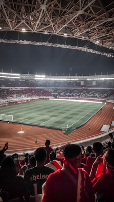 A large stadium filled with spectators, predominantly dressed in red, overlooks a well-lit football field with distinct grass patterns. The stadium's architecture features an intricate roof design with exposed beams. The field is surrounded by a running track, and the atmosphere appears lively and anticipatory.