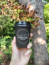 A hand is holding a black coffee cup with the logo 'coffee-bike' and 'since 2016' printed on it. The background features lush green foliage and a tree trunk, suggesting an outdoor, natural setting.