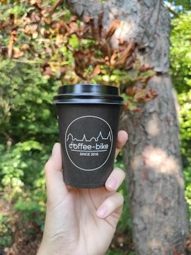 A hand is holding a black coffee cup with the logo 'coffee-bike' and 'since 2016' printed on it. The background features lush green foliage and a tree trunk, suggesting an outdoor, natural setting.