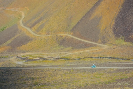 A winding dirt road stretches across a barren landscape with rolling hills and sparse vegetation. A single turquoise car travels along the road, emphasizing the vastness of the area. The terrain is rugged with earthy tones, and minimal vegetation can be seen.