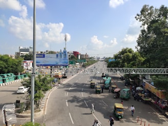A busy urban street scene featuring multiple lanes of traffic, including cars and auto rickshaws. A large billboard is visible on the left, and commercial buildings are present in the background. The road is flanked by trees and there is a pedestrian bridge spanning across it. People are visible walking along the street.