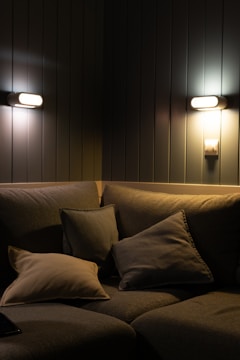 A cozy corner with a fabric sofa adorned with multiple soft cushions, all in a muted palette. Two warm wall lights illuminate the space against a backdrop of dark, vertical paneling.