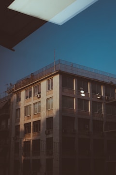 A multi-story building with large windows and air conditioning units on the exterior. The angle of the photograph captures the building from below, and a reflection is visible in the upper part, showing a portion of the ceiling or roof lighting within a different structure.