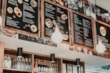 A stylish eatery features large wooden-framed menu boards hanging overhead, displaying a variety of dishes and drinks, including salads, laksa, nasi lemak, sides, drinks, and desserts. Below, shelves are stocked with assorted bottled beverages and condiments. Ambient lighting from hanging lights adds warmth to the scene.