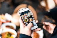 A person holding a smartphone capturing a photo of several colorful dishes on a table. The image focuses on the phone screen displaying the food, with blurred hands and food in the background. The scene is vibrant and contrasts natural with electronic elements.