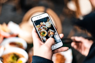 A person holding a smartphone capturing a photo of several colorful dishes on a table. The image focuses on the phone screen displaying the food, with blurred hands and food in the background. The scene is vibrant and contrasts natural with electronic elements.