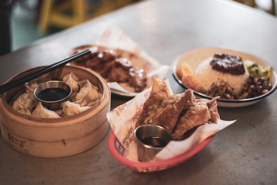 A tabletop features a variety of Asian-inspired dishes. In the foreground, there is a bamboo steamer filled with dumplings accompanied by a small container of soy sauce and chopsticks resting on the steamer. Nearby, a serving of fried wontons is presented in a small red basket lined with parchment paper, alongside another container of dipping sauce. In the background, a bowl of rice topped with a dark sauce is served with various side accompaniments like greens and beans.