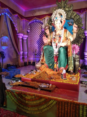 A detailed and ornate statue of Lord Ganesha is seated on an intricately designed throne, surrounded by floral decorations and offerings. The background features draped curtains and columns, with colorful lighting enhancing the festive atmosphere.