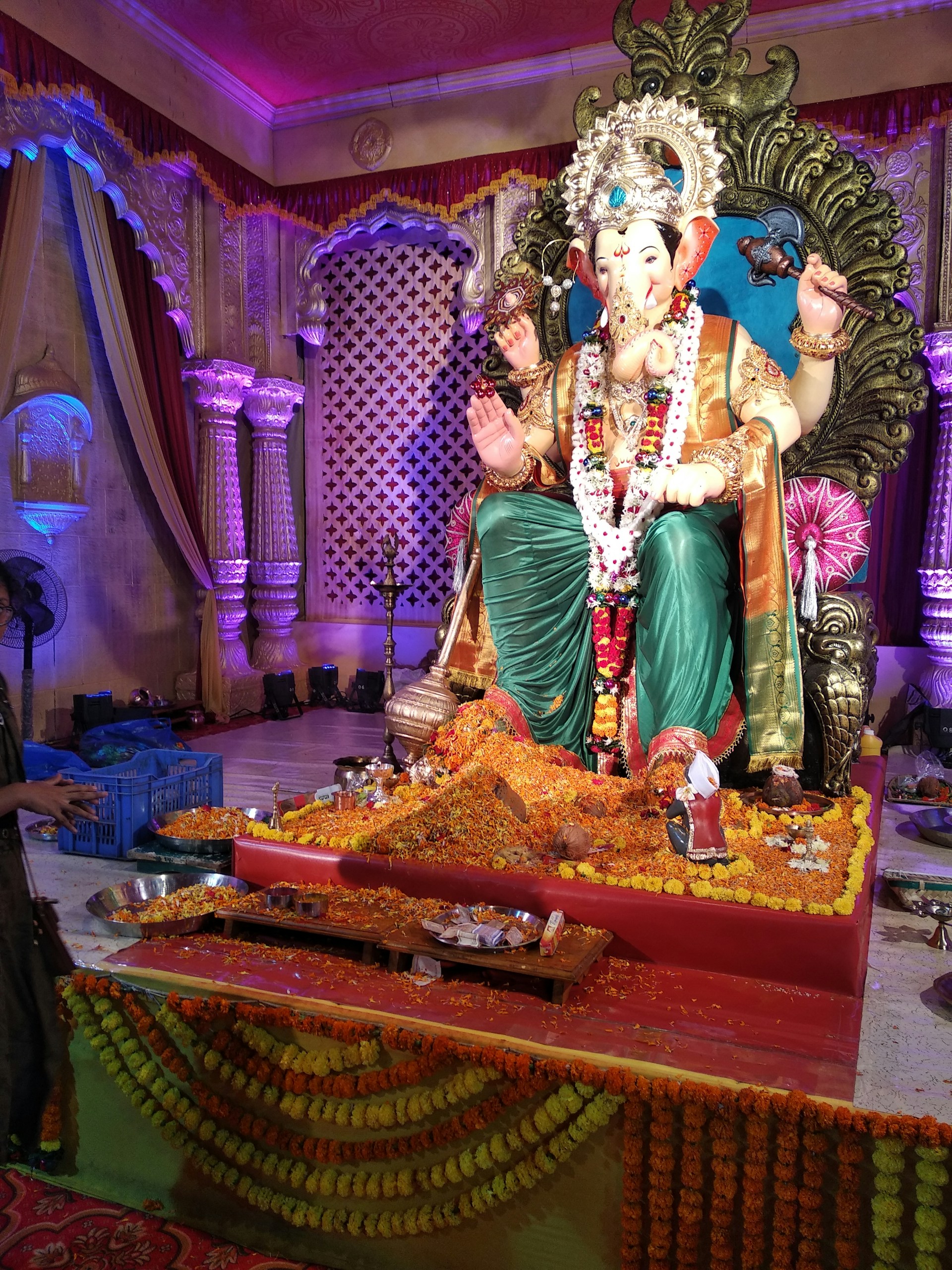 A detailed and ornate statue of Lord Ganesha is seated on an intricately designed throne, surrounded by floral decorations and offerings. The background features draped curtains and columns, with colorful lighting enhancing the festive atmosphere.