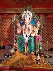 A large, intricately decorated statue of the Hindu deity Ganesha is seated on an ornate platform. The statue is adorned with colorful garlands, jewels, and a majestic headdress. In front of the statue, there are flower petals and offerings. The background features an elaborately designed stage with warm lighting.