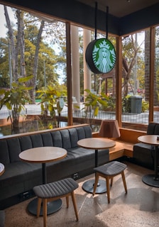 A cozy interior of a coffee shop with wooden tables and stools, featuring a couch alongside large windows. Outside, greenery is visible through the glass. The shop's emblem is suspended from the ceiling, and the environment is brightened by natural light.