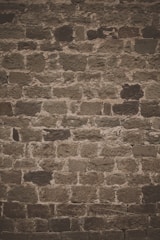 A textured stone wall composed of irregularly shaped, roughly hewn stones with varying shades of brown and gray. The stones are set in a pattern that forms a rustic and weathered appearance.