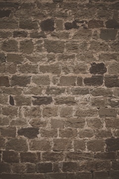 A textured stone wall composed of irregularly shaped, roughly hewn stones with varying shades of brown and gray. The stones are set in a pattern that forms a rustic and weathered appearance.