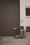 A black barbecue grill on wheels, positioned on a concrete surface in front of a brown, closed garage door. The grill has a rectangular shape with side handles and a lower shelf. The surrounding area features a white wall and a small patch of grass is visible at the bottom left corner.