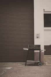 A black barbecue grill on wheels, positioned on a concrete surface in front of a brown, closed garage door. The grill has a rectangular shape with side handles and a lower shelf. The surrounding area features a white wall and a small patch of grass is visible at the bottom left corner.