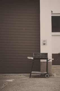 A black barbecue grill on wheels, positioned on a concrete surface in front of a brown, closed garage door. The grill has a rectangular shape with side handles and a lower shelf. The surrounding area features a white wall and a small patch of grass is visible at the bottom left corner.