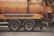 A side view of an industrial vehicle with large wheels, featuring a bright orange cylindrical tank, likely used for cleaning or transporting liquids. The vehicle is parked on a grey asphalt surface and some traffic cones are visible in the background.
