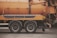 A side view of an industrial vehicle with large wheels, featuring a bright orange cylindrical tank, likely used for cleaning or transporting liquids. The vehicle is parked on a grey asphalt surface and some traffic cones are visible in the background.