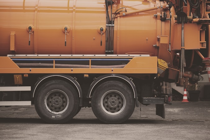 A side view of an industrial vehicle with large wheels, featuring a bright orange cylindrical tank, likely used for cleaning or transporting liquids. The vehicle is parked on a grey asphalt surface and some traffic cones are visible in the background.