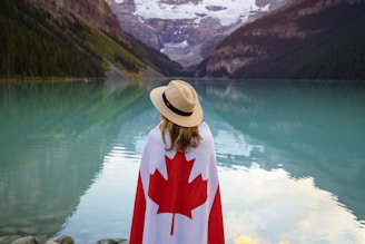 A person draped in a Canadian flag stands by a serene, turquoise lake surrounded by forested mountains and snow-capped peaks. The individual is wearing a straw hat and looking towards the picturesque view.