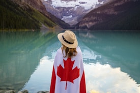 A person draped in a Canadian flag stands by a serene, turquoise lake surrounded by forested mountains and snow-capped peaks. The individual is wearing a straw hat and looking towards the picturesque view.