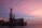 An offshore drilling rig is situated on a calm body of water, under a sky painted with pink and purple hues from the setting sun. The structure is industrial and complex, with tall towers and various machinery visible.