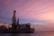 An offshore drilling rig is situated on a calm body of water, under a sky painted with pink and purple hues from the setting sun. The structure is industrial and complex, with tall towers and various machinery visible.
