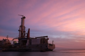 An offshore drilling rig is situated on a calm body of water, under a sky painted with pink and purple hues from the setting sun. The structure is industrial and complex, with tall towers and various machinery visible.