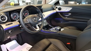 The interior of a high-end car showcasing a sleek design with luxurious black leather seats and a modern dashboard. The dashboard and control panel feature advanced technology displays and a distinct Mercedes-Benz logo on the steering wheel. Ambient lighting casts a subtle blue glow along the contours of the interior, enhancing the sophisticated atmosphere. The materials used include wood and metallic finishes, contributing to the opulent aesthetic.