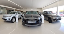 A showroom with several Land Rover vehicles displayed prominently on a glossy tiled floor. The vehicles include a white SUV, a black Discovery Sport, and a silver SUV. Bright natural light streams in through large windows, illuminating the area.