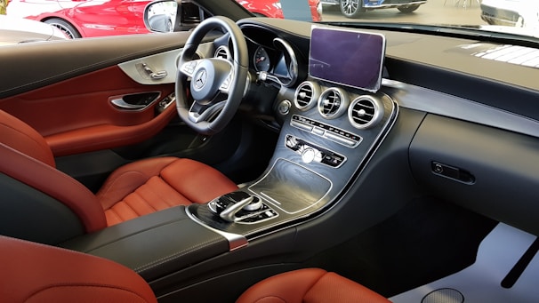 The interior of a luxury car featuring red leather seats and a sleek dashboard with modern controls. The steering wheel bears an iconic logo, and the center console has a large display screen and various buttons. The surrounding interior is primarily black with metallic accents.