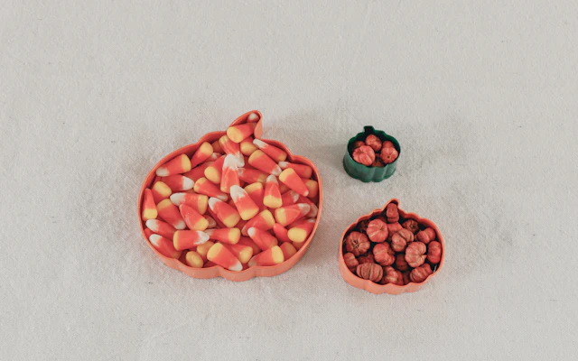 A pumpkin-shaped dish of candy corn beside two smaller dishes on a pale surface