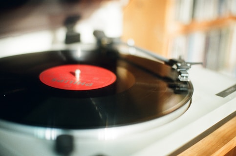 A close-up of a vinyl record on a turntable with a red label in the center. The turntable's needle is positioned on the record, suggesting music is either playing or about to play. The background is softly blurred, likely to emphasize the focus on the record player.