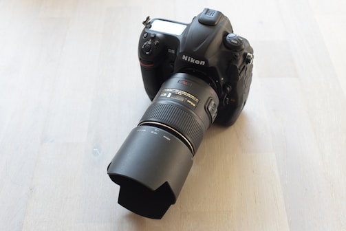 A professional Nikon DSLR camera with a large telephoto lens is positioned on a light wooden surface. The camera appears robust and high-tech, featuring detailed buttons and a digital display on the back.