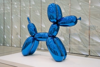A large, shiny blue balloon sculpture shaped like a dog is displayed on a white platform. The reflective surface of the sculpture distorts the surrounding environment, creating an abstract and playful visual effect. The background features a modern, architectural wall with geometric patterns.