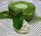 A circular green tea mousse cake is garnished with a mint leaf and a dollop of whipped cream, plated on a dish with a swirling black and white pattern. Matcha powder is lightly dusted over the cake and the plate.