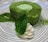 A circular green tea mousse cake is garnished with a mint leaf and a dollop of whipped cream, plated on a dish with a swirling black and white pattern. Matcha powder is lightly dusted over the cake and the plate.