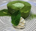 A circular green tea mousse cake is garnished with a mint leaf and a dollop of whipped cream, plated on a dish with a swirling black and white pattern. Matcha powder is lightly dusted over the cake and the plate.