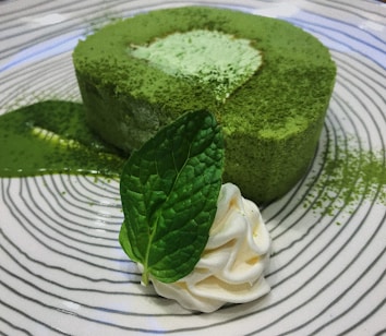 A circular green tea mousse cake is garnished with a mint leaf and a dollop of whipped cream, plated on a dish with a swirling black and white pattern. Matcha powder is lightly dusted over the cake and the plate.