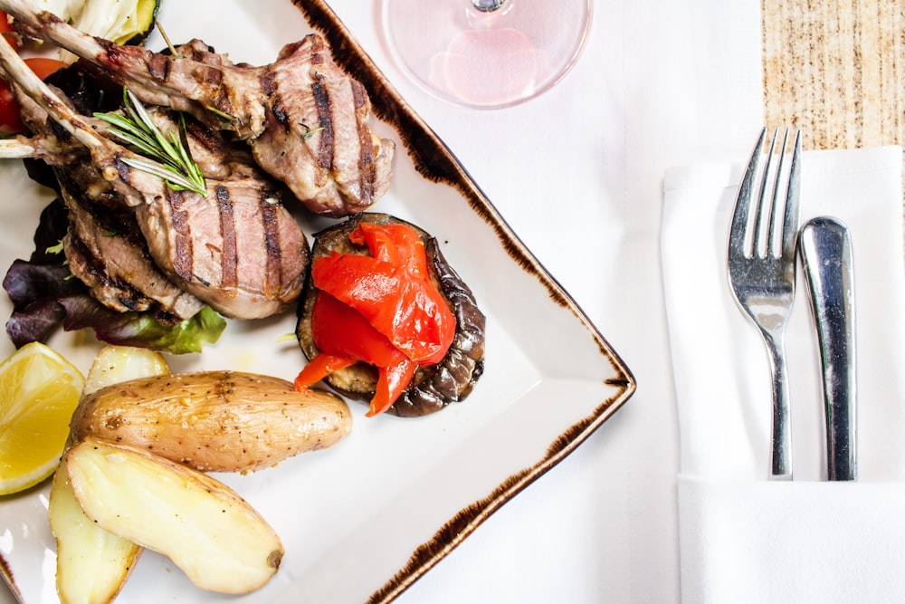 well-done steak near sliced potato in plate near gray stainless steel fork