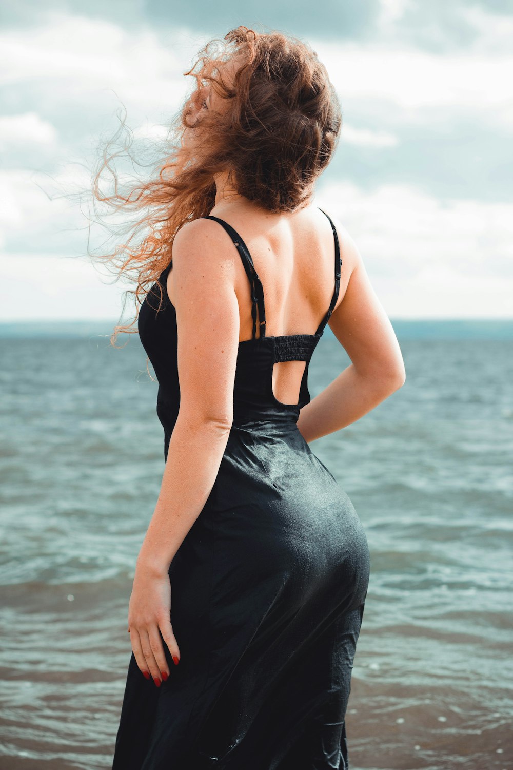 立っていると昼間の間に青い海に面した黒いスパゲッティストラップドレスを着ている女性