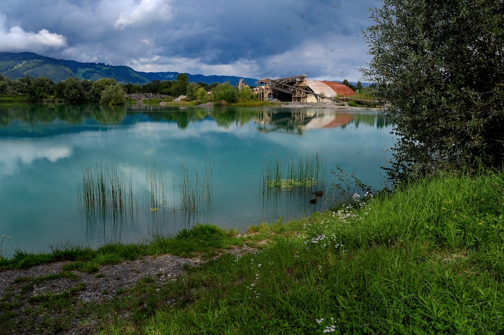 house near lake surrounded with tall and green trees viewing mountain under blue and white skies during daytime