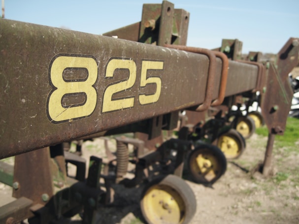 A close-up view of a piece of agricultural machinery featuring the number 825 in large yellow lettering. The machine appears to be old and worn, with rust visible on the metal. The background shows several wheels and parts of the machinery, indicating it's possibly a planter or similar farm equipment.