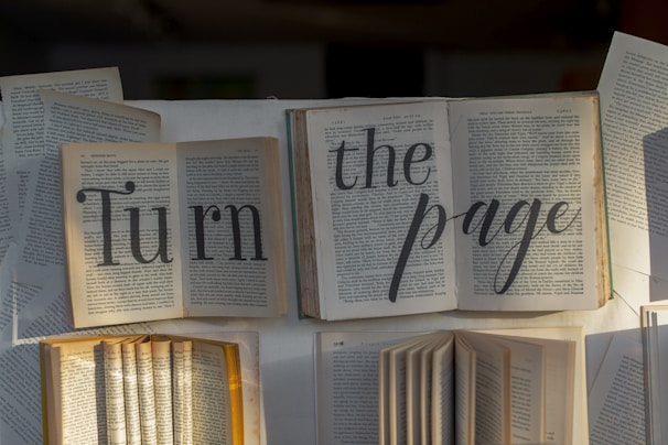 Several open books are arranged in such a way that the words 'Turn the page' are clearly visible. The pages of the books are gently fanned out, and natural light casts soft shadows across the text, highlighting the message.